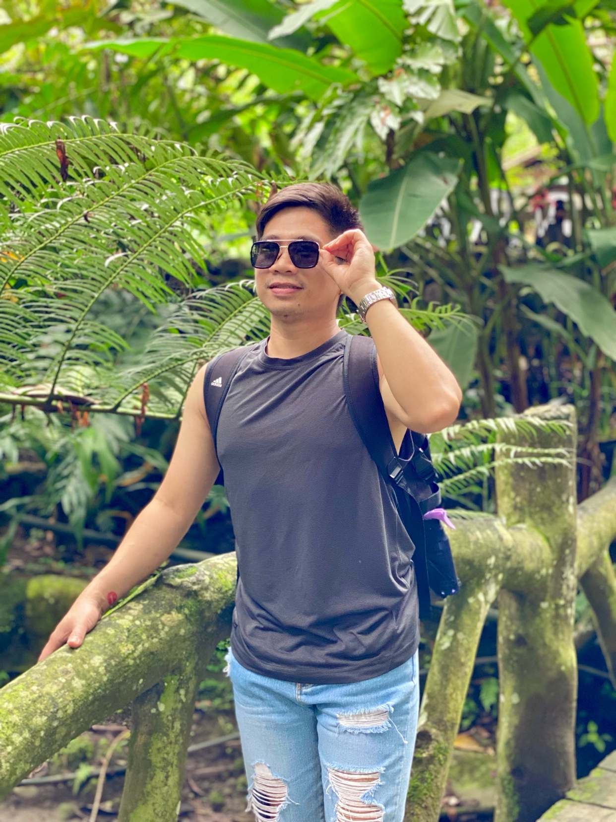 A man poses for a photo in a lush, green outdoor setting. He wears sunglasses and a casual outfit, smiling at the camera.