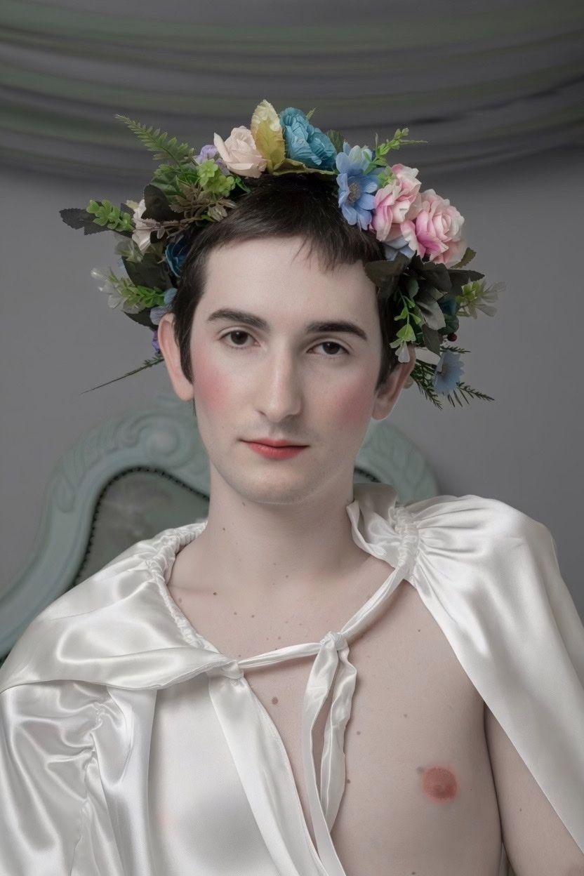 A person with short dark hair and a flower crown sits in front of a grey backdrop. They are wearing a white satin cape and makeup, including blush and red lipstick.