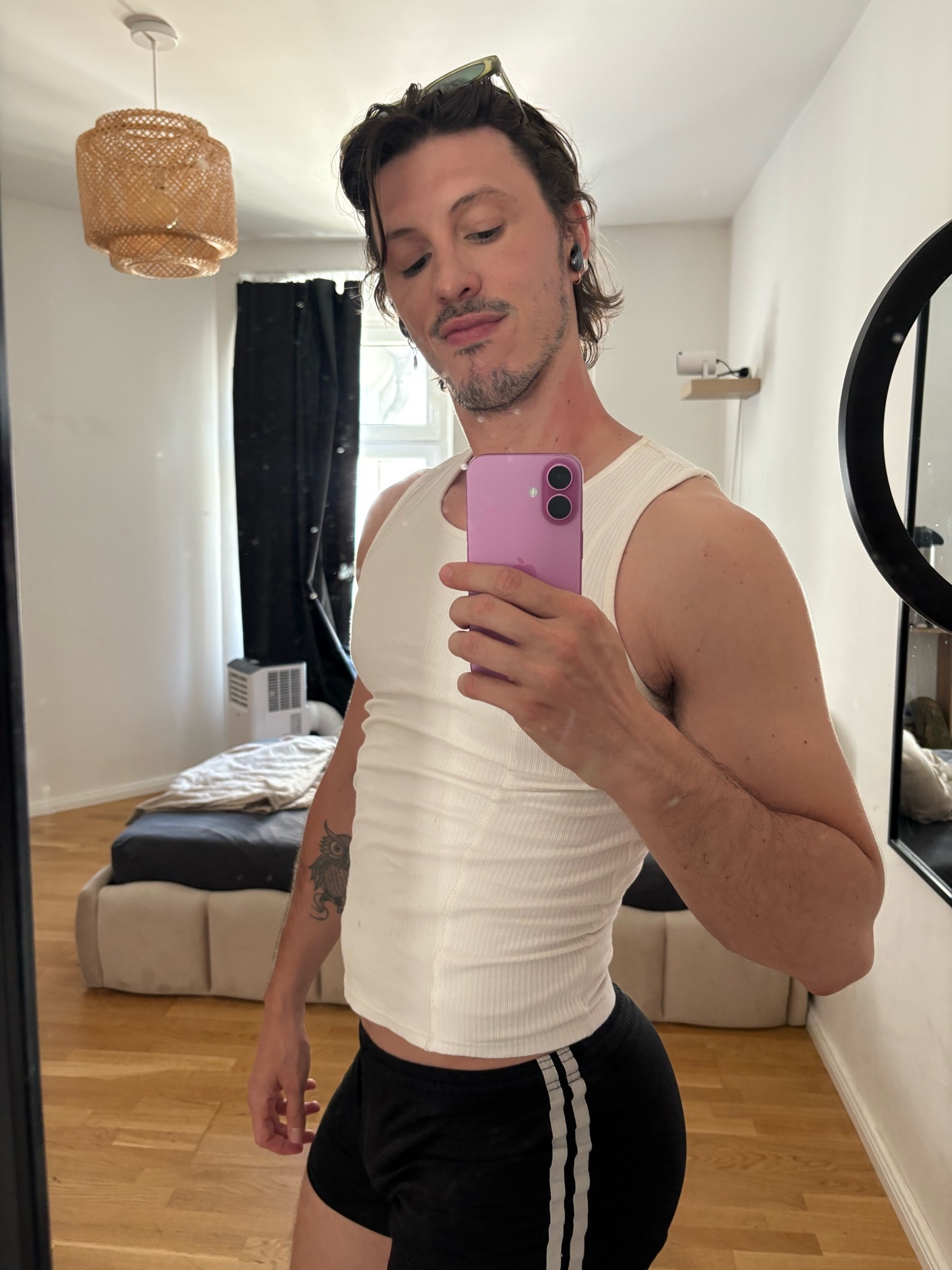 A man in a white tank top and black shorts takes a mirror selfie in a bedroom. He is wearing sunglasses on his head and has tattoos on his arm.