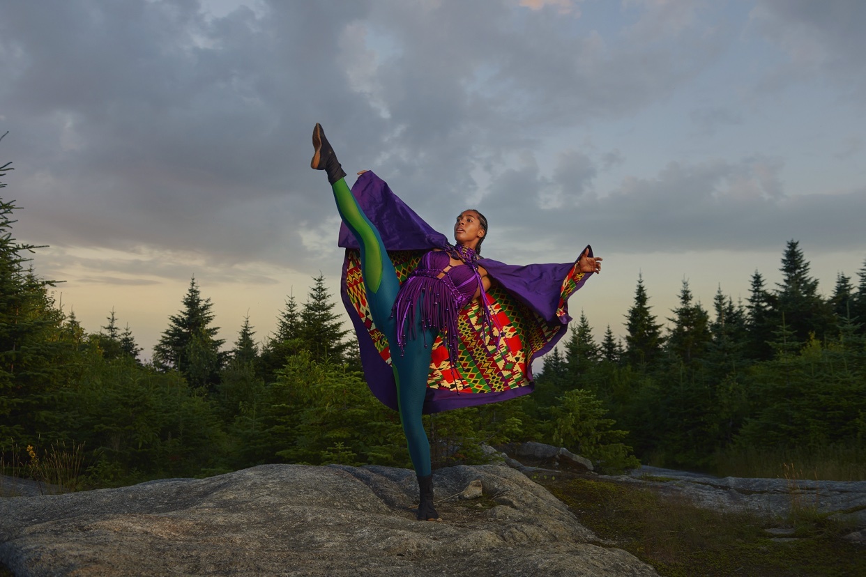 A woman in a vibrant purple cape and green leggings performs a high leg kick while balancing on a rock in a forest clearing. The dramatic sky above suggests a sense of movement and possibility.