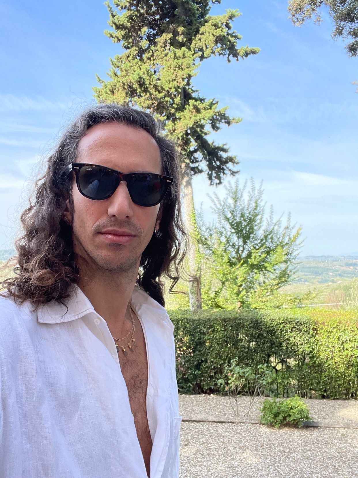 A man with long, curly hair and sunglasses takes a selfie outdoors. He is wearing a white shirt and jewelry.
