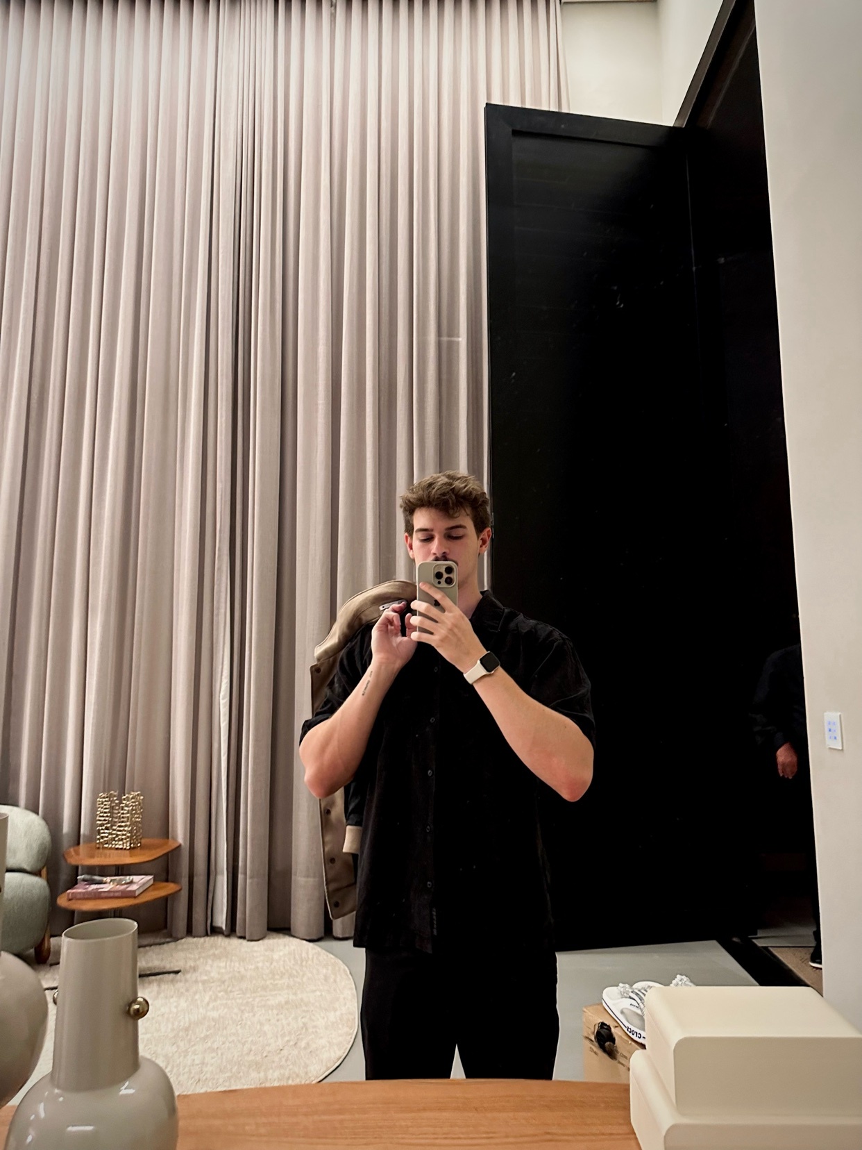 A man takes a mirror selfie in a modern, well-lit room. He is wearing a black button-down shirt and holding a jacket over his shoulder.