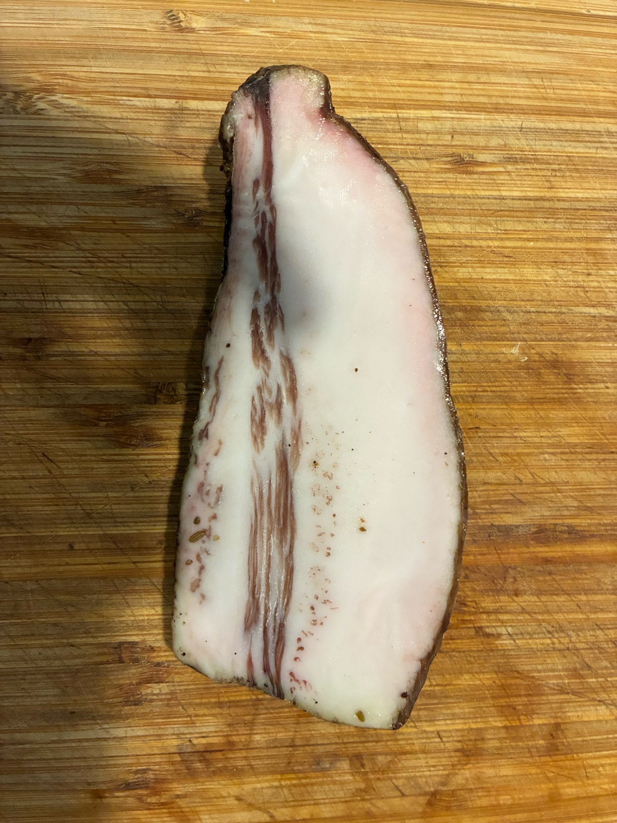 A close-up, overhead shot of a single slice of homemade guanciale, a cured pork product, resting on a wooden cutting board. The guanciale is thick and has distinct layers of white fat and reddish-brown meat, with some darker speckles.
