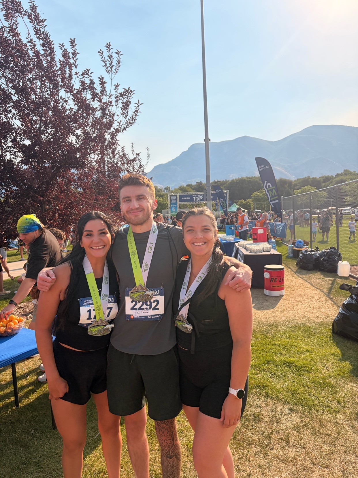 Three friends pose for a photo wearing race medals around their necks and bibs on their shirts. The man in the middle has his arms around the two women on either side of him. They are all smiling and appear to have just finished a race, with a crowd