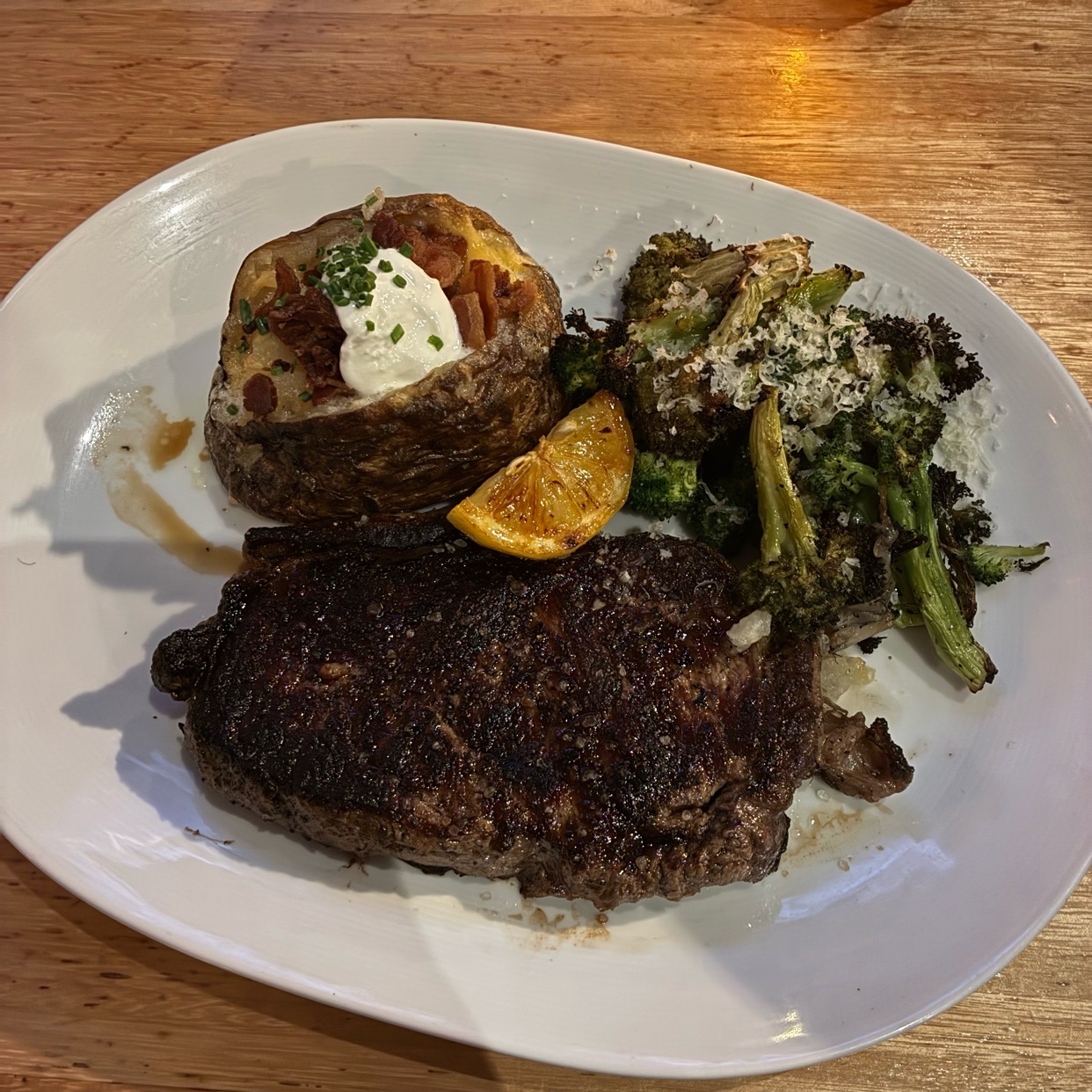 A close-up shot of a steak dinner on a white plate. The meal includes a steak, a loaded baked potato, and roasted broccoli. The steak is dark brown and appears to be grilled. The baked potato is cut in half and topped with sour cream, chives, and bac