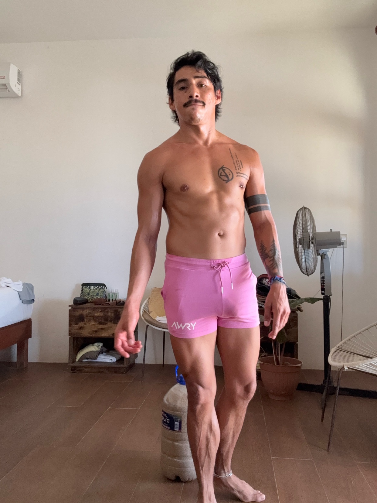 A man poses shirtless, showcasing his muscular physique. He wears pink shorts and has tattoos on his arm.