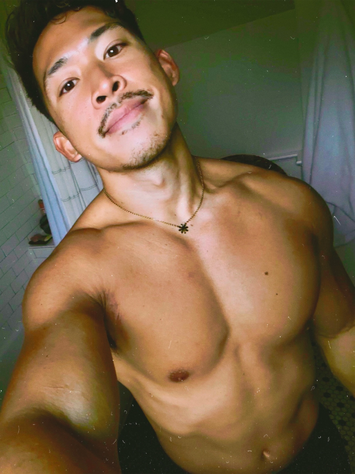 A man takes a selfie in a bathroom, showcasing his muscular physique. He is shirtless, with a mustache and a necklace.