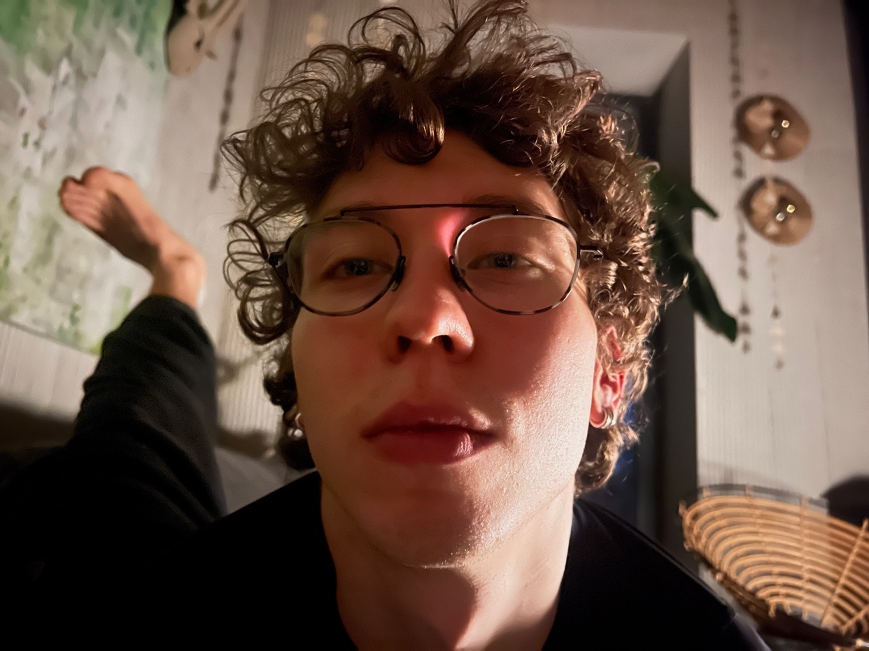 A selfie of a man with curly hair and glasses. His leg is visible in the background.
