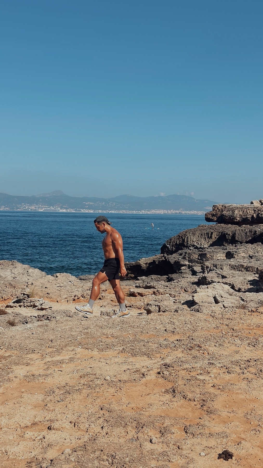 A shirtless, muscular man walks along a rocky coastline with the ocean in the background under a clear blue sky.