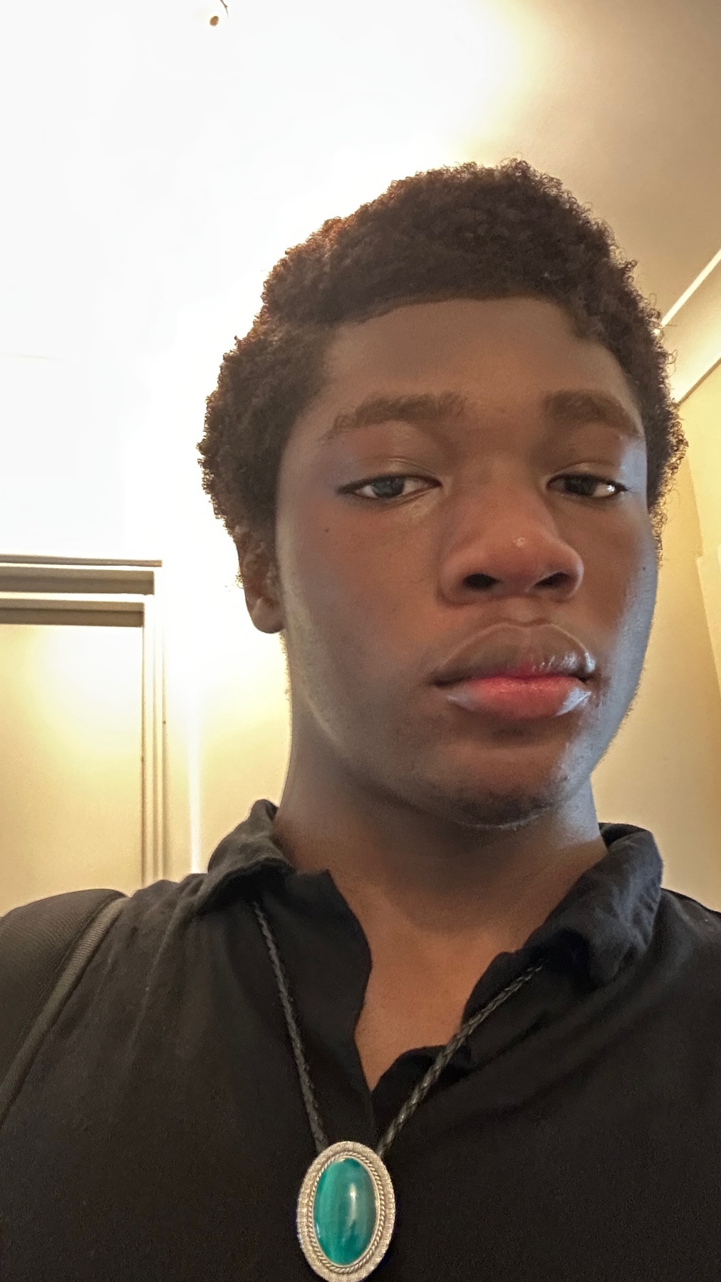 A close-up selfie of a young black man with curly hair and a serious expression. He is wearing a black collared shirt and a necklace with a large turquoise pendant.
