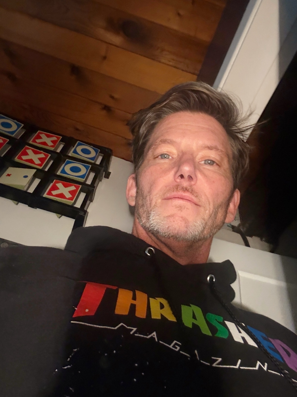 A man with stubble and a rainbow logo hoodie poses for a selfie in front of a tic tac toe game.