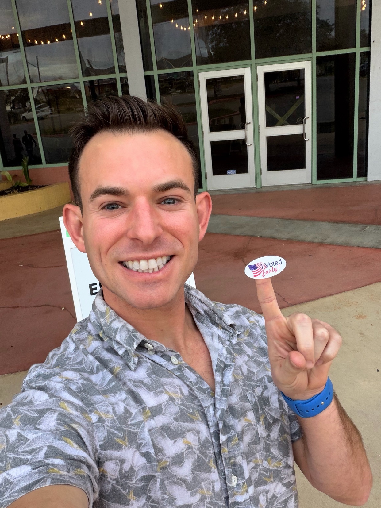 A man smiles at the camera while holding up an "I Voted" sticker on his finger. He is wearing a grey and white patterned short-sleeved shirt and standing outside a building with large glass doors.