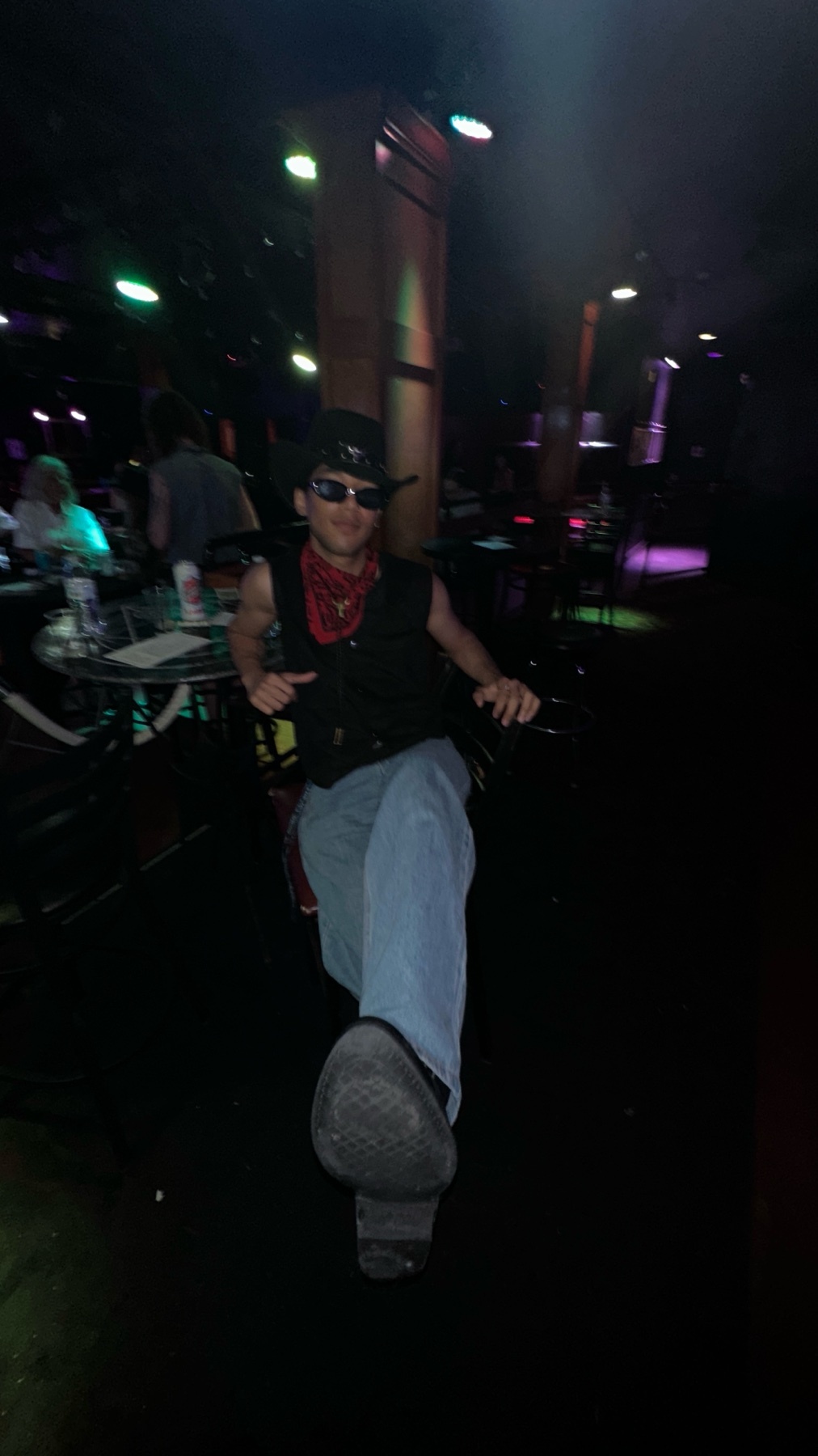 A man in a cowboy hat, sunglasses, bandana, vest, and jeans poses in a dimly lit nightclub.