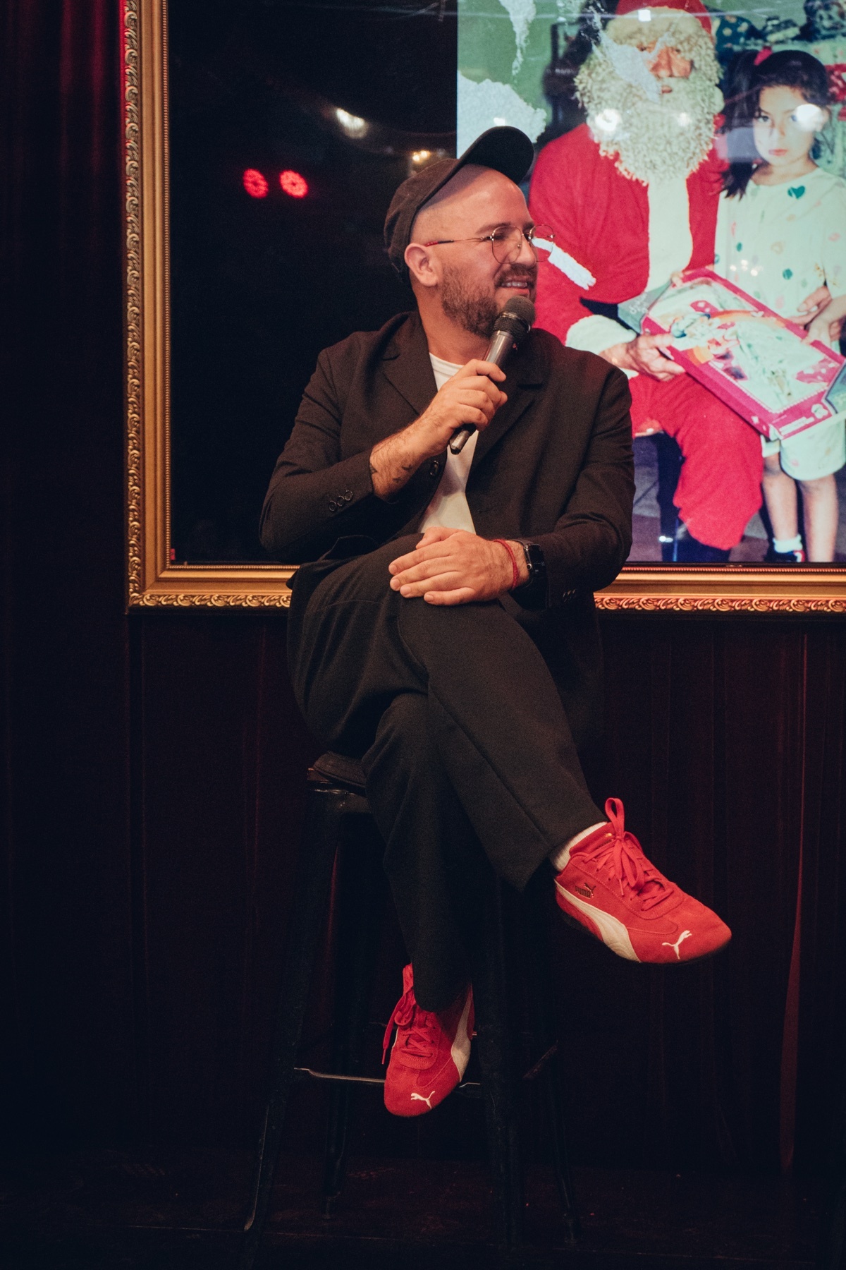 A man with a bald head and a beard is sitting on a stool, holding a microphone. He is wearing a suit and red shoes.