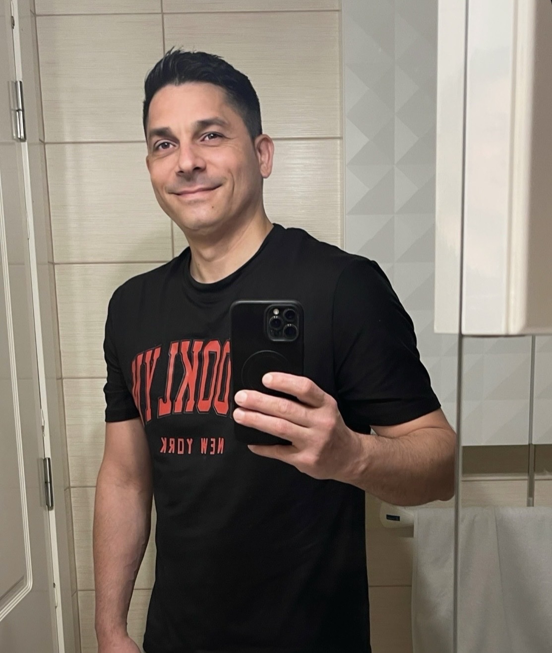 A man in a black t-shirt with "New York" written on it takes a selfie in a bathroom.