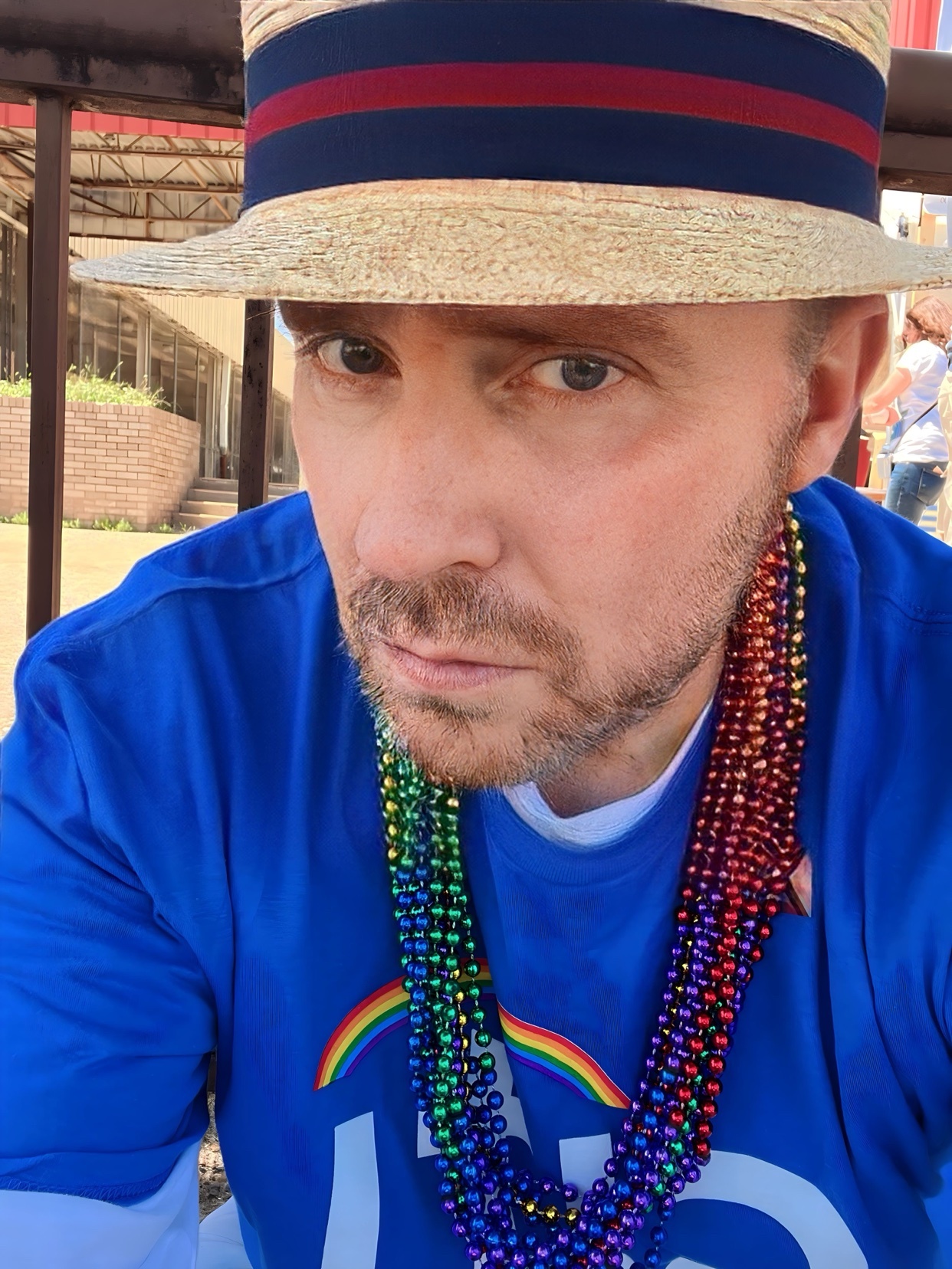 A man wearing a straw hat and a blue shirt with rainbow beads around his neck poses for a close-up selfie.
