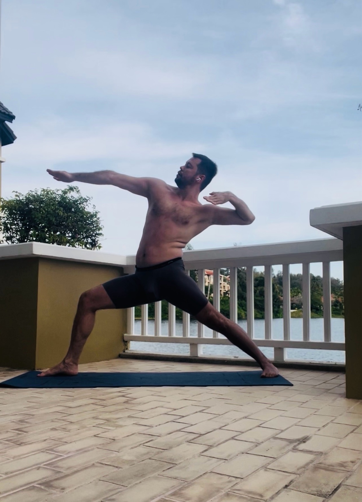 A man is doing yoga on a mat outside. He is shirtless and in a warrior pose.