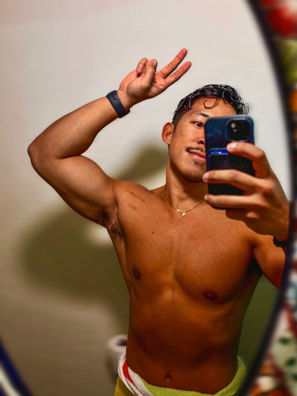 A man takes a mirror selfie, flexing his arm and making a peace sign. He is shirtless, showing off his muscular physique.