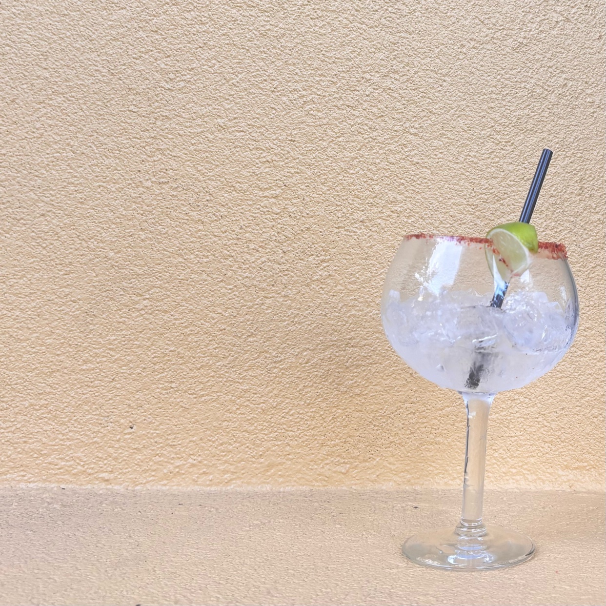 A refreshing cocktail with ice, lime, and a salt rim sits in a large glass against a textured yellow wall.