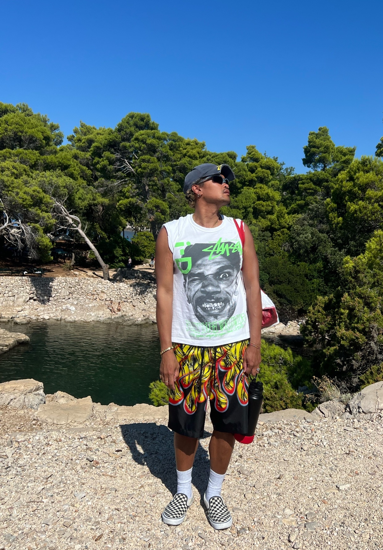 A man stands outdoors wearing a graphic t-shirt, flame-patterned shorts, a baseball cap, and sunglasses. He is accessorized with a backpack and a water bottle, posing in front of a scenic backdrop of trees and water under a clear blue sky.