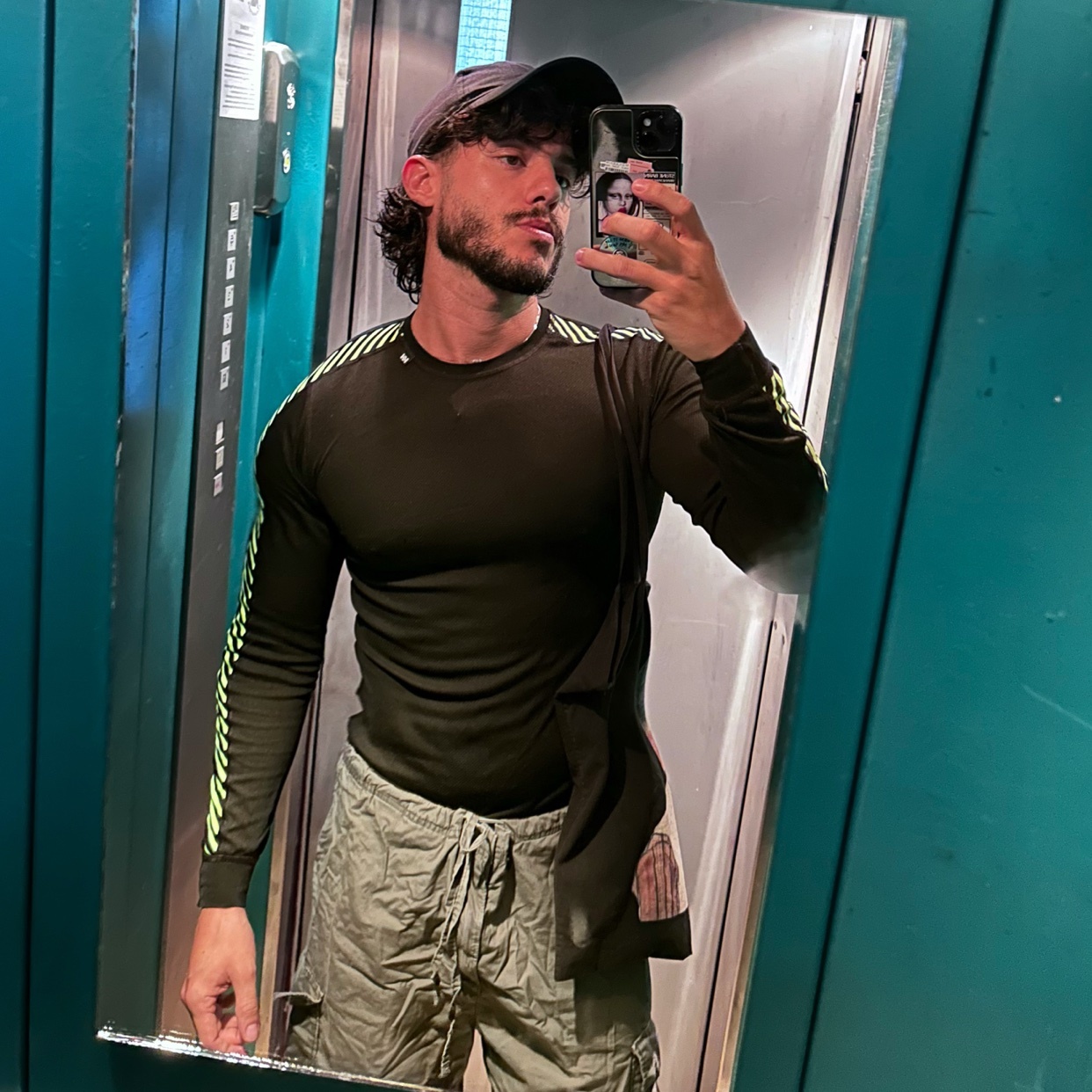 A man takes a mirror selfie, showcasing his physique. He's wearing a cap and a long-sleeved shirt, with a towel draped over his shoulder.