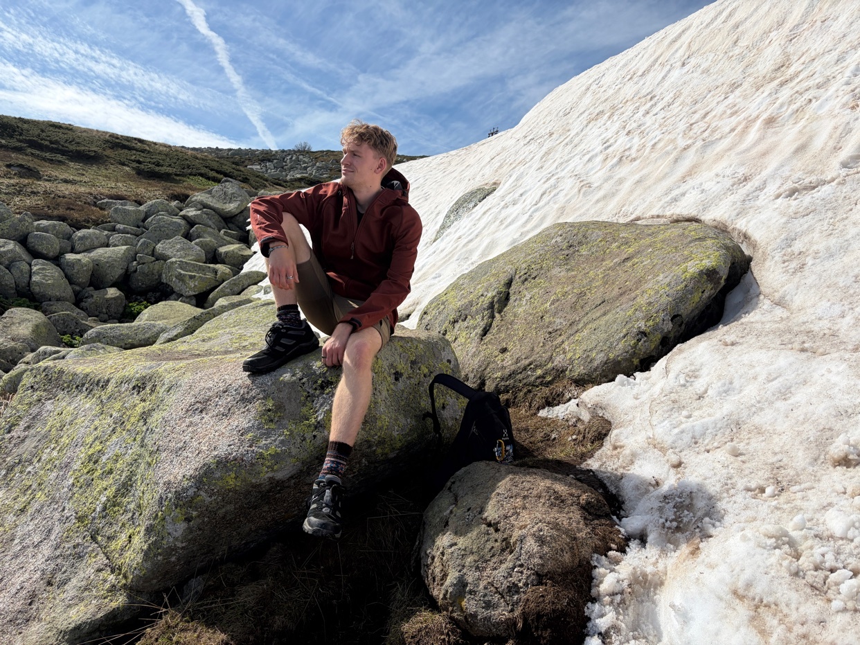 A young man sits on large rocks in a snowy mountain landscape, looking off to the side. He is wearing a maroon jacket, shorts, and hiking boots, with a backpack resting beside him.