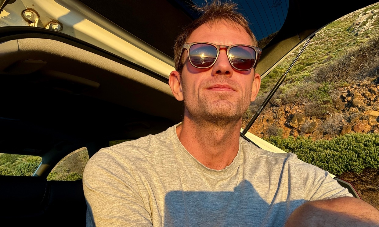 A man wearing sunglasses and a t-shirt poses for a selfie in the back of a car during golden hour. The sun reflects off his sunglasses, and the warm light illuminates the rocky, outdoor landscape behind him.