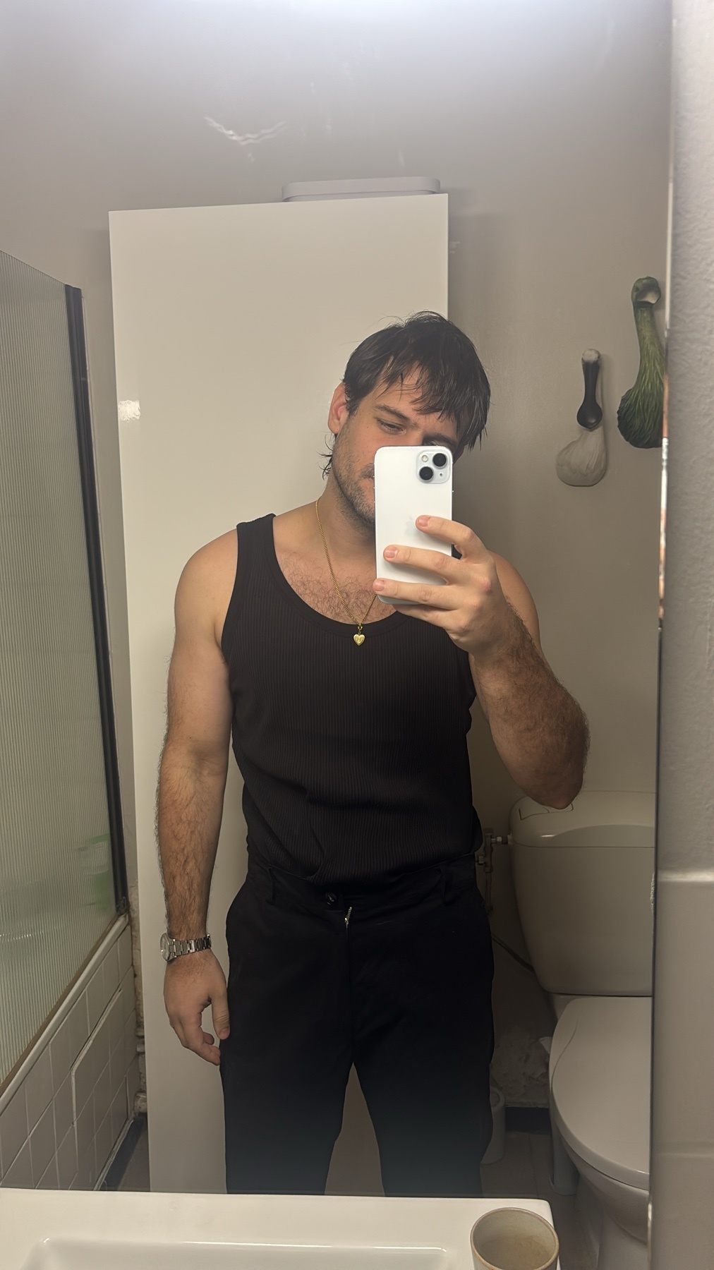 A man takes a mirror selfie in a bathroom. He is shirtless and wearing a black tank top and black pants.