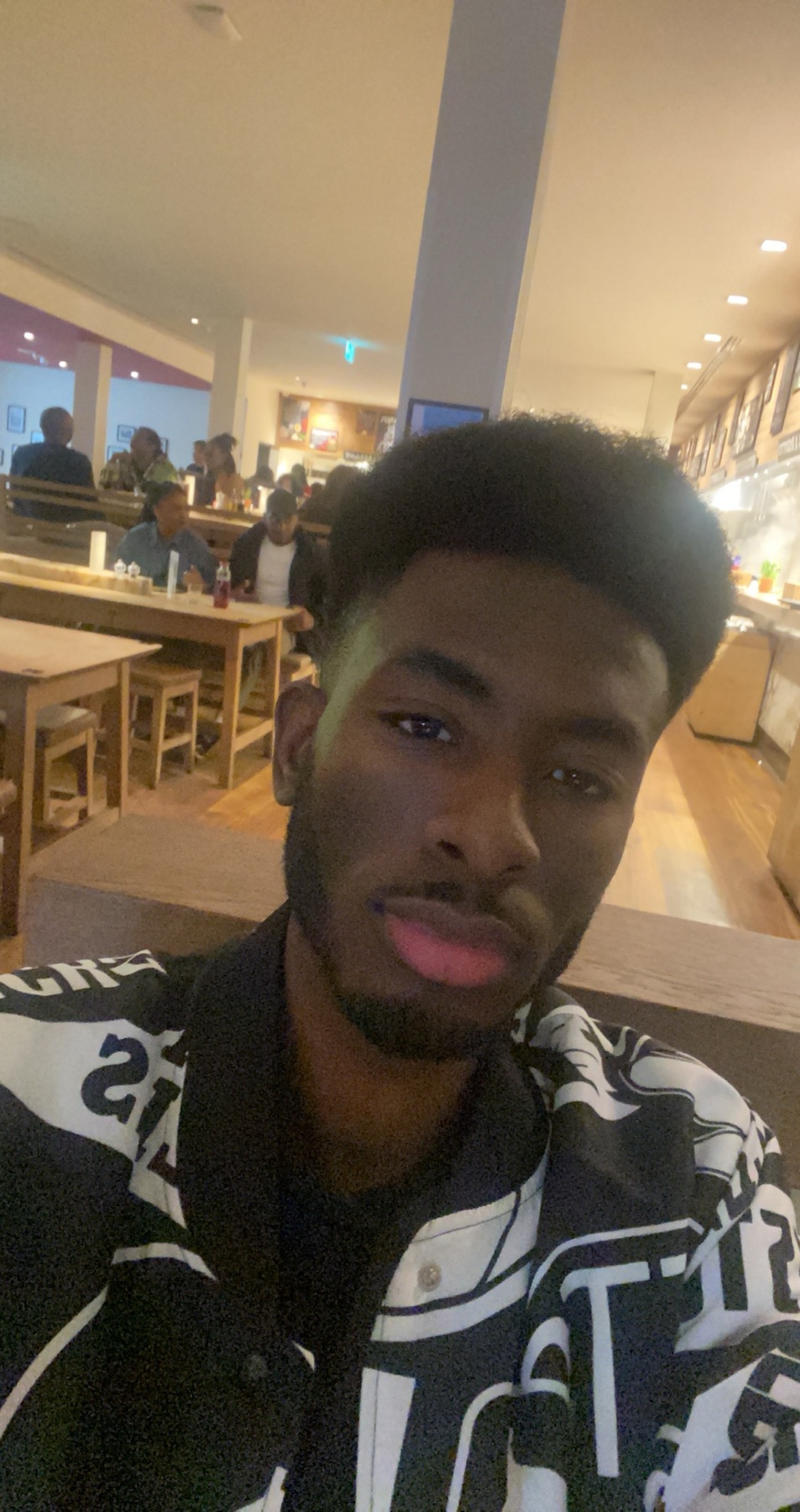 A man with a beard and dark hair takes a selfie in a restaurant. He is wearing a black and white patterned jacket.