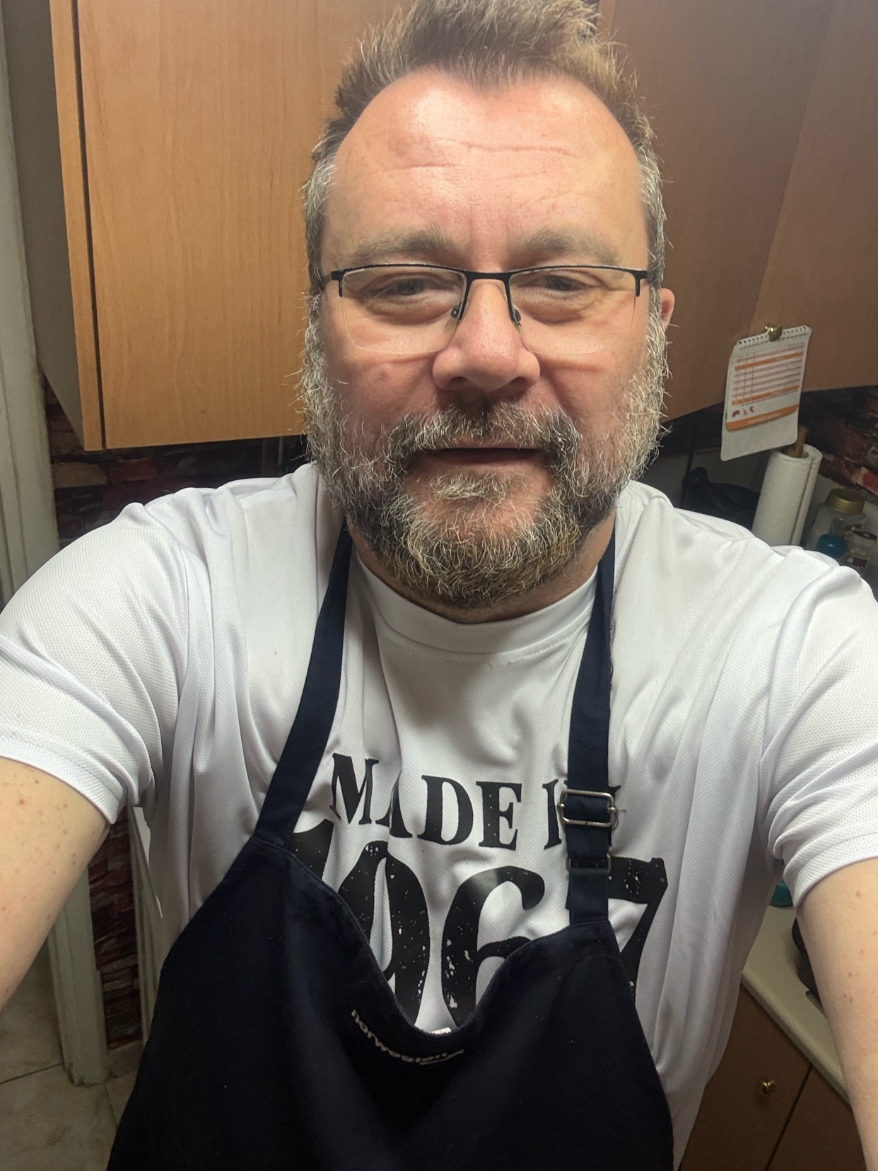 A man with a beard and glasses wears a white t-shirt and a dark apron with the words "MADE IN 1967" printed on it. He is in a kitchen setting and appears to be taking a selfie.