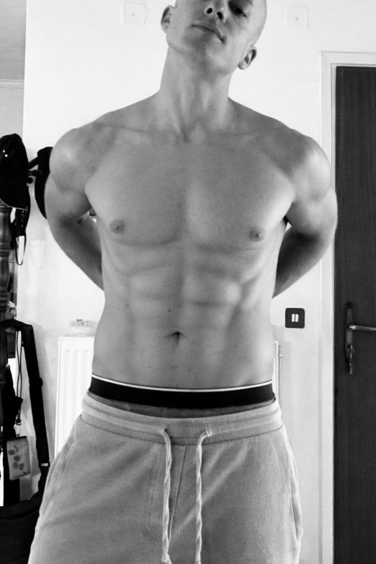 A black and white, medium shot of a man's torso. He is shirtless, wearing sweatpants, and has visible abs and muscular arms. His head is tilted back and his hands are behind his back.