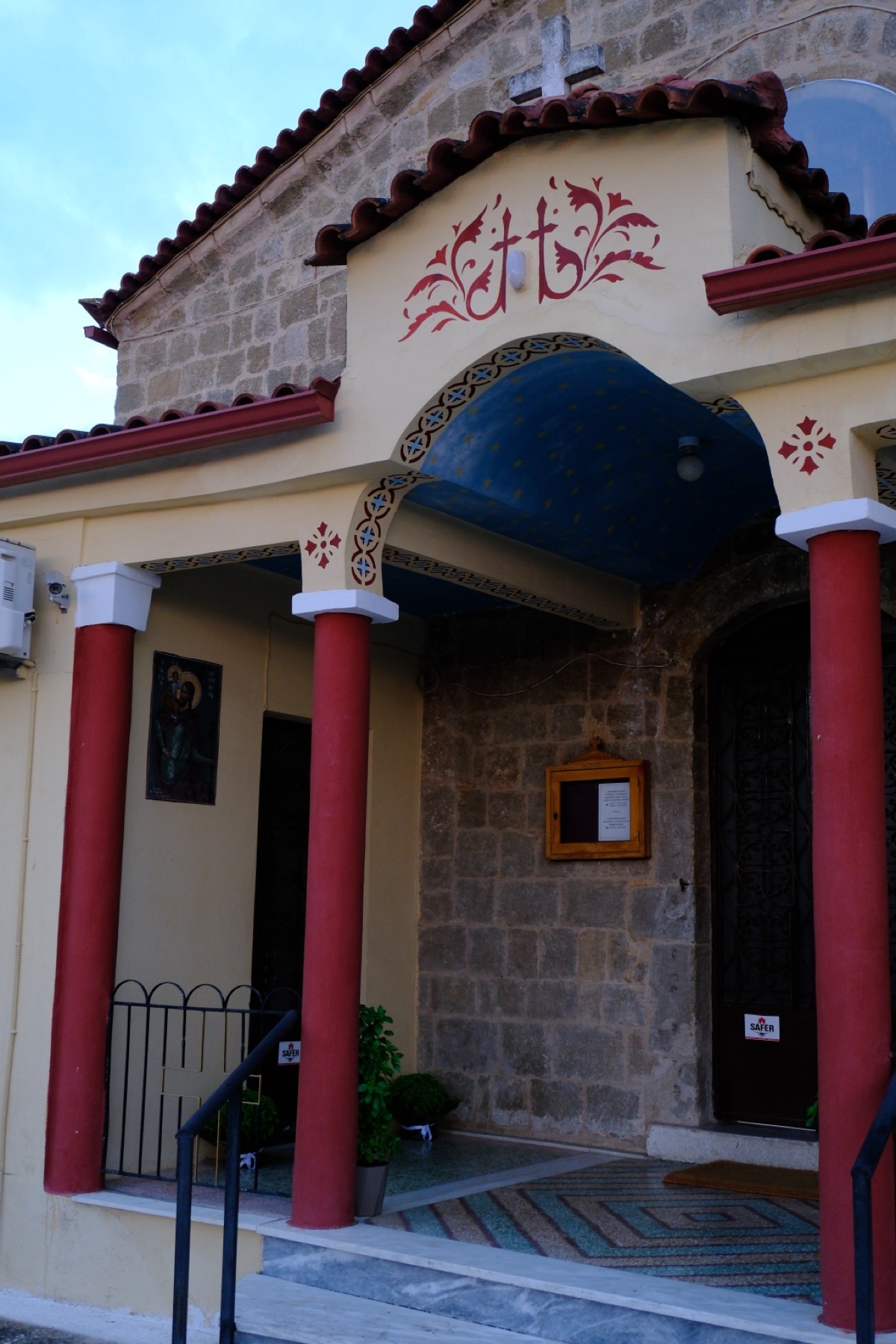 The entrance to a beautiful Greek Orthodox church features red columns, a blue ceiling, and intricate decorative crosses above the arched doorway. The stone facade and tiled roof add to its charming architectural details.