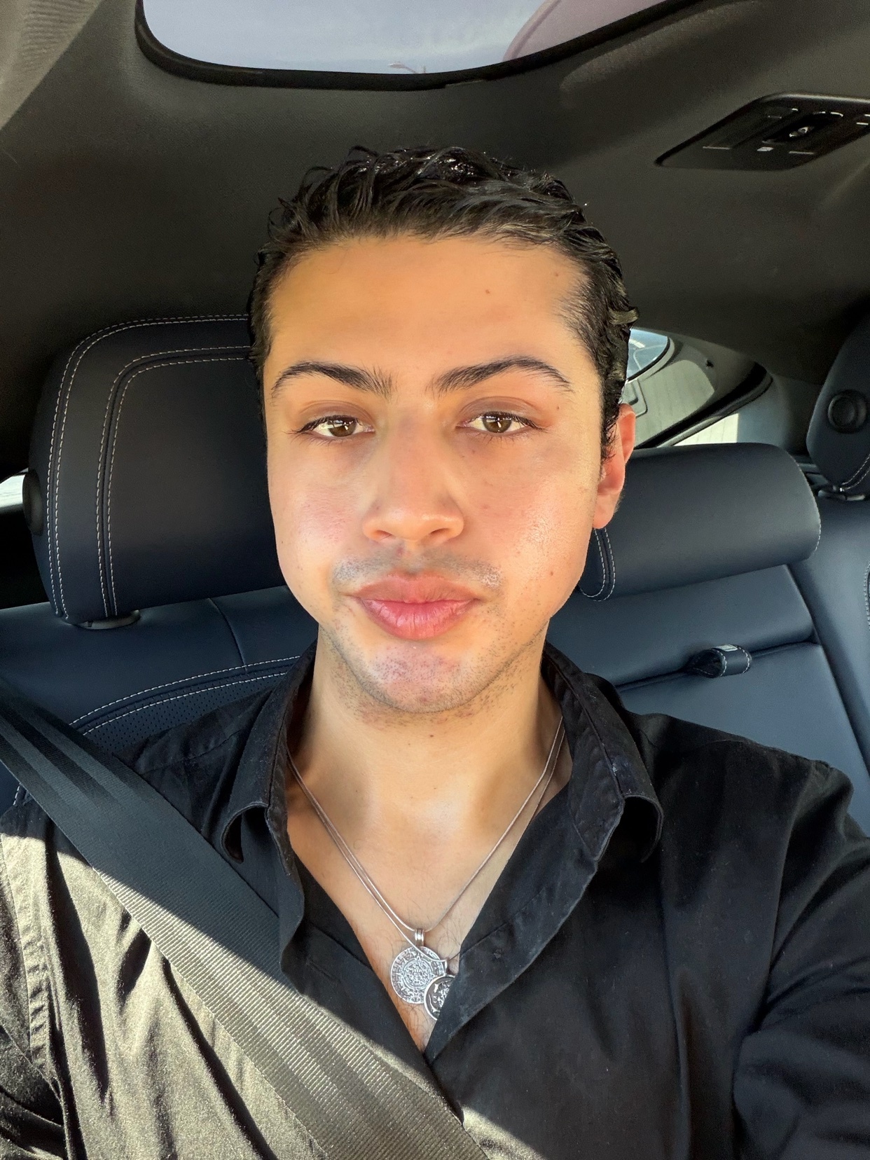 A man with dark, slicked-back hair and a black collared shirt takes a selfie in the driver's seat of a car.