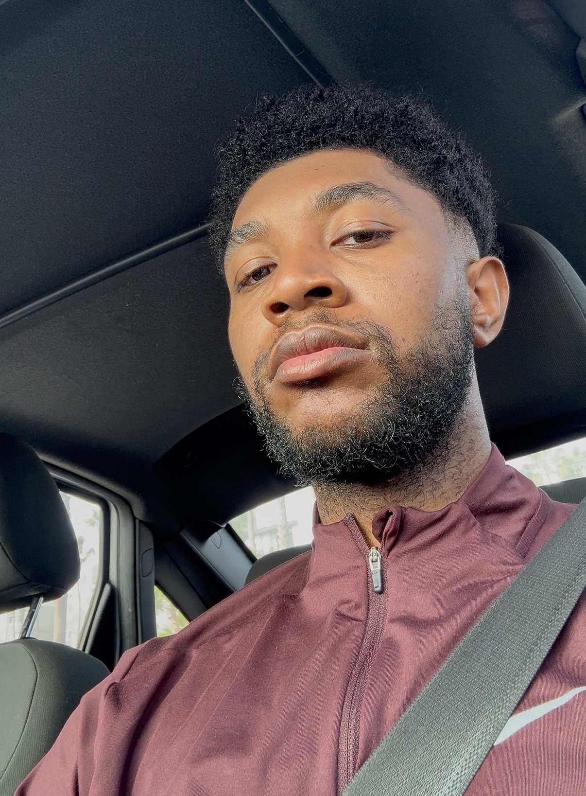 A man takes a selfie in a car. He has dark hair and a beard.