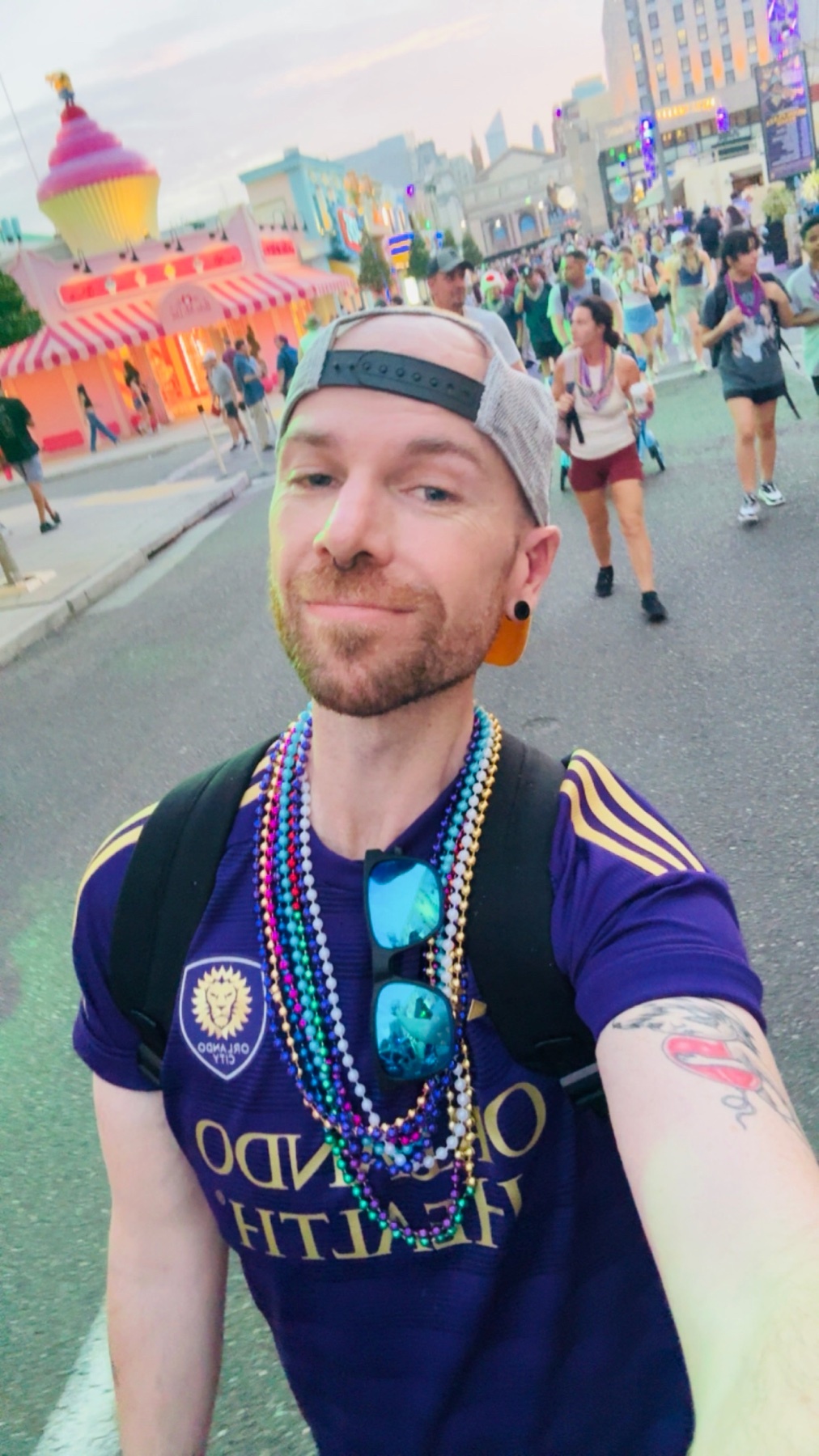 A man wearing a purple Orlando City SC jersey and a grey baseball cap takes a selfie in front of a bustling street scene. He is adorned with multiple strands of colorful beads around his neck and has sunglasses tucked into his shirt.
