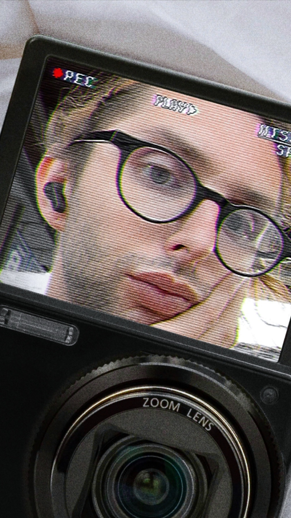 A close-up selfie of a man wearing round glasses and earbuds, captured through the lens of a vintage camera. The man appears to be in a somber mood, with his hand resting on his cheek.