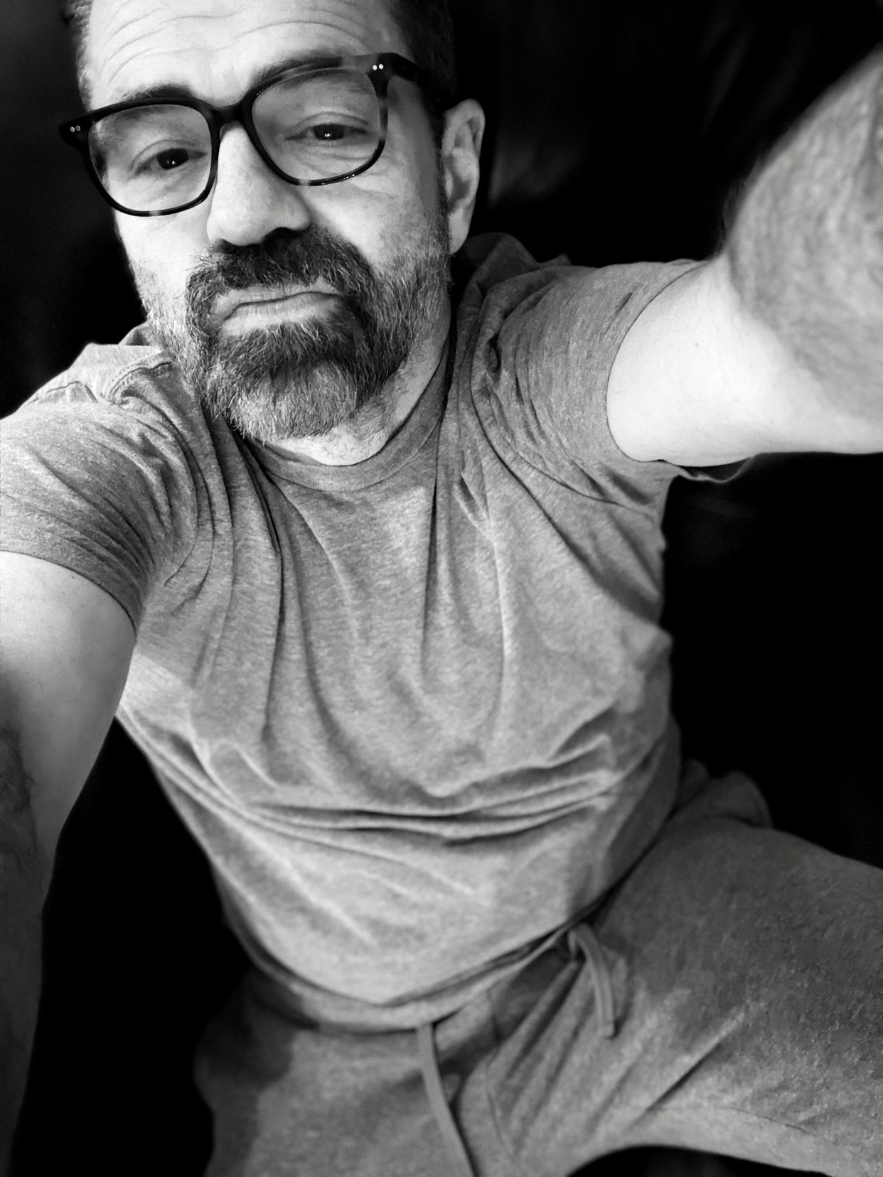 A black and white selfie of a man wearing glasses and a t-shirt. The man has a beard and appears to be indoors.