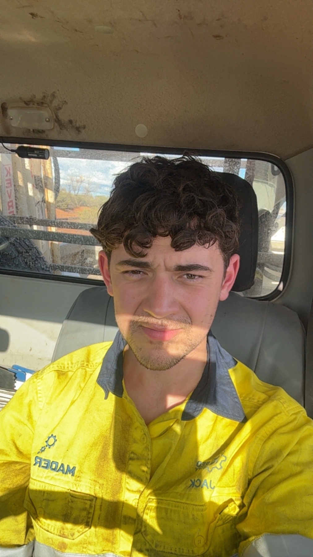 A man with curly dark hair and a light mustache poses for a selfie in the driver's seat of a vehicle. He is wearing a bright yellow work shirt with blue accents on the collar and sleeves.