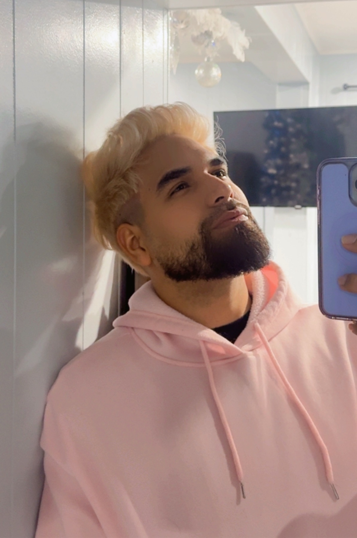 A man with blonde hair and a beard takes a mirror selfie. He is wearing a pink hoodie.