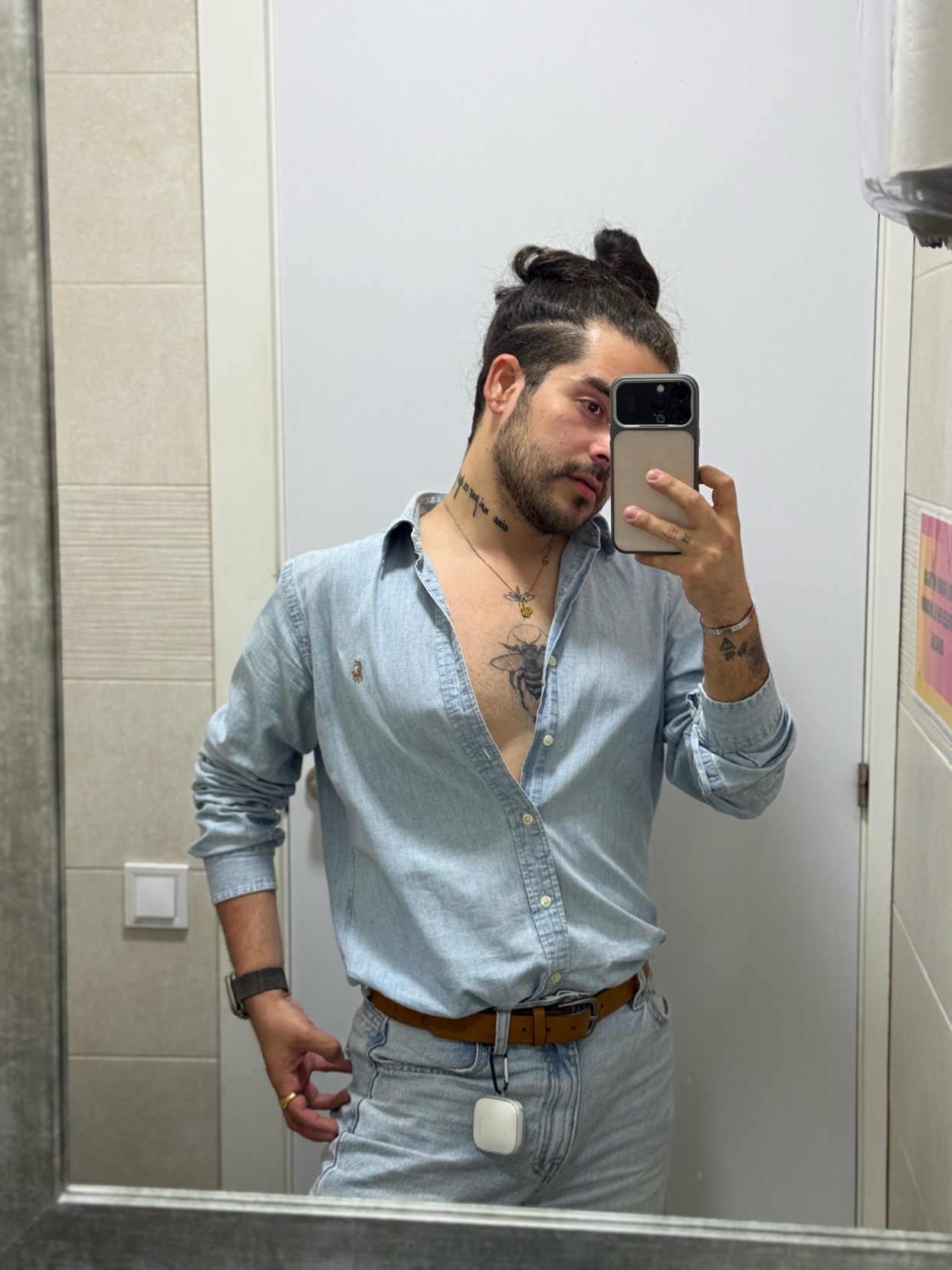 A man with a man bun and tattoos takes a mirror selfie in a light blue denim shirt and jeans.