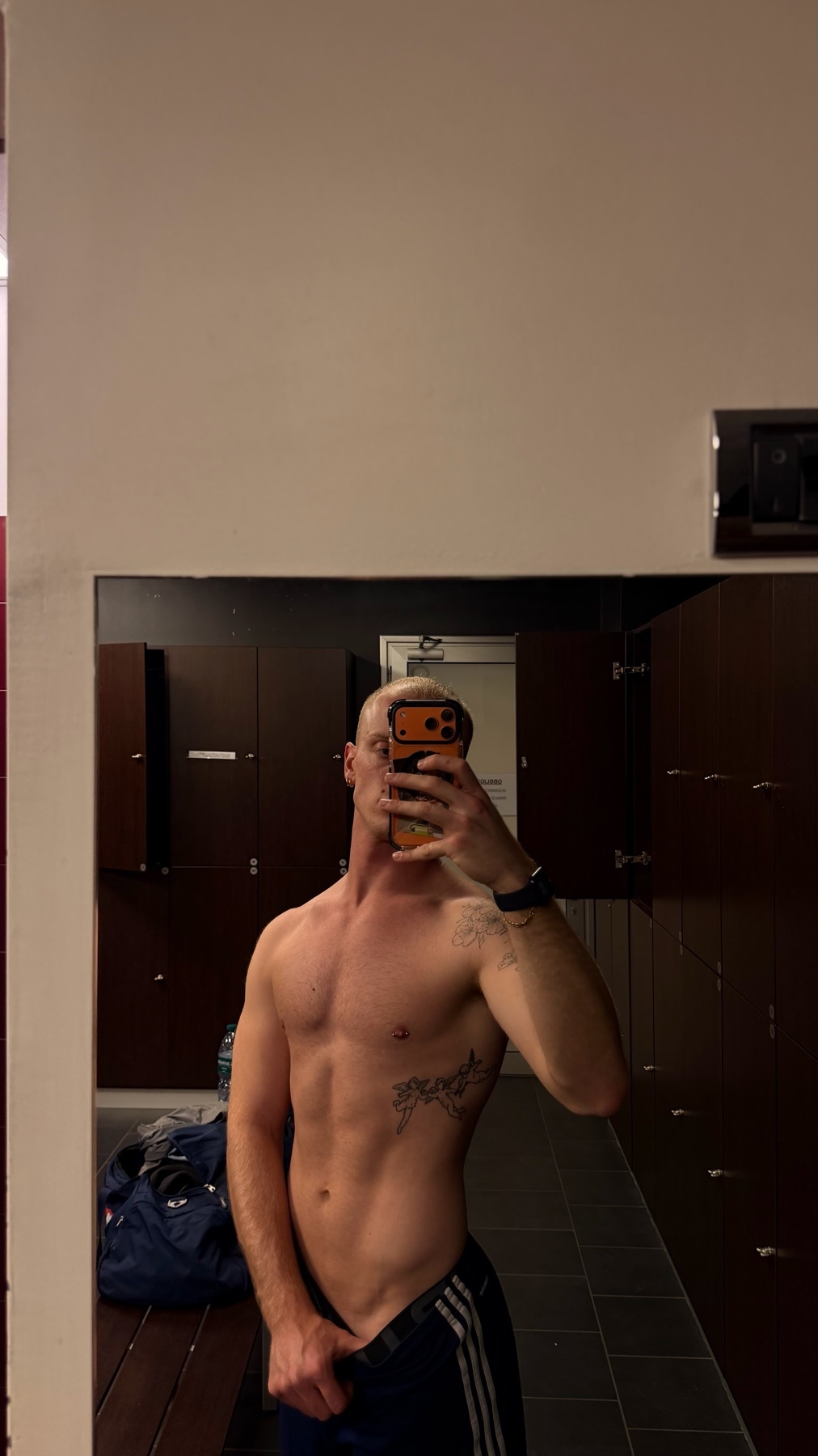 A shirtless man with tattoos takes a mirror selfie in a locker room. He is flexing his abs and showing off his muscular physique.