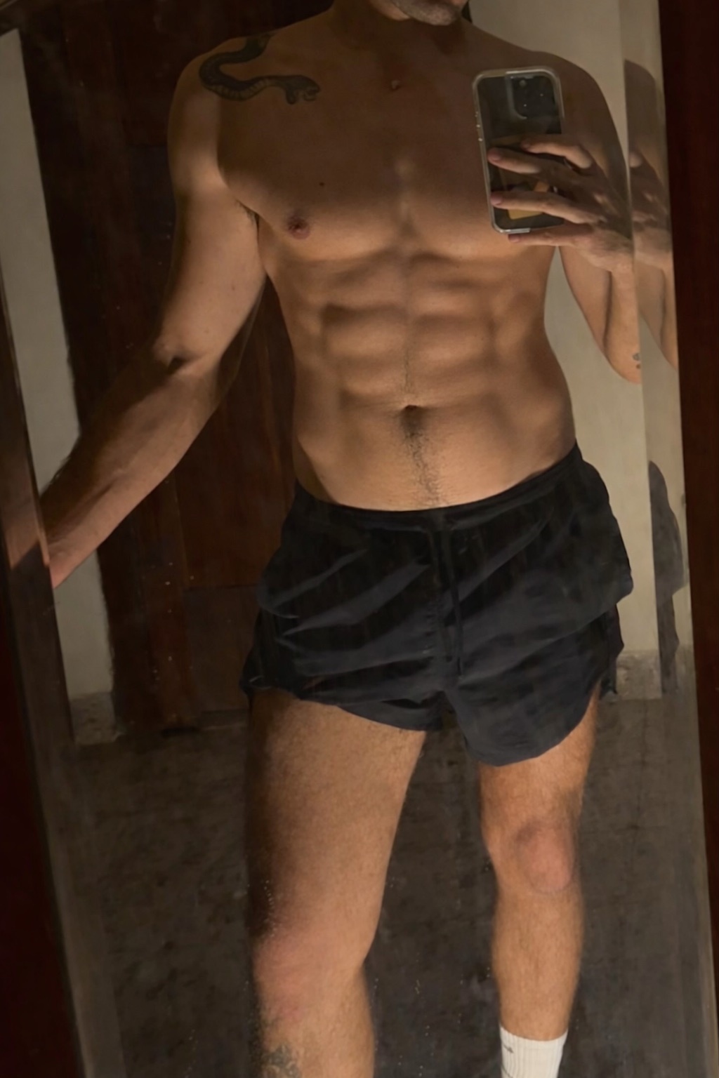 A man takes a mirror selfie, showing off his muscular physique and abs while wearing black shorts and white socks. He has tattoos on his arm and leg.