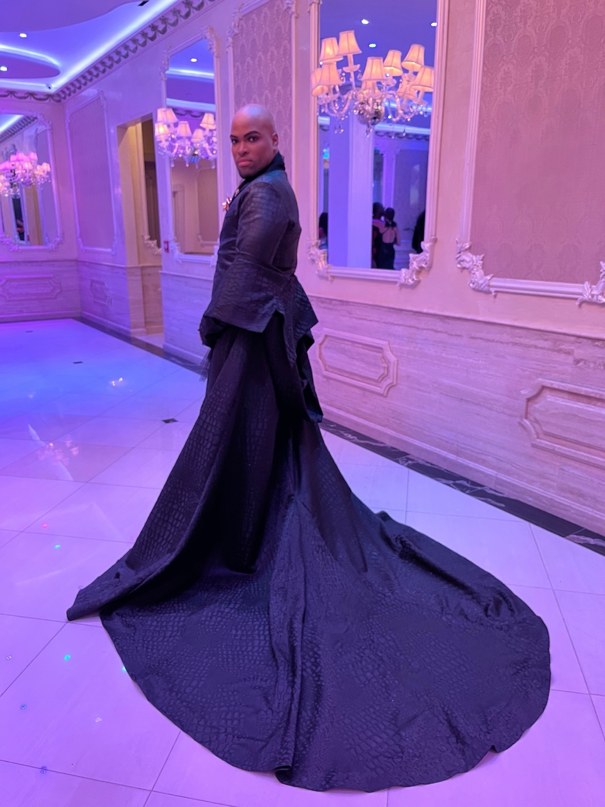 A man in a black dress with a long train poses in a room with ornate mirrors and chandeliers. The photo is taken from behind, showcasing the dress's dramatic design.