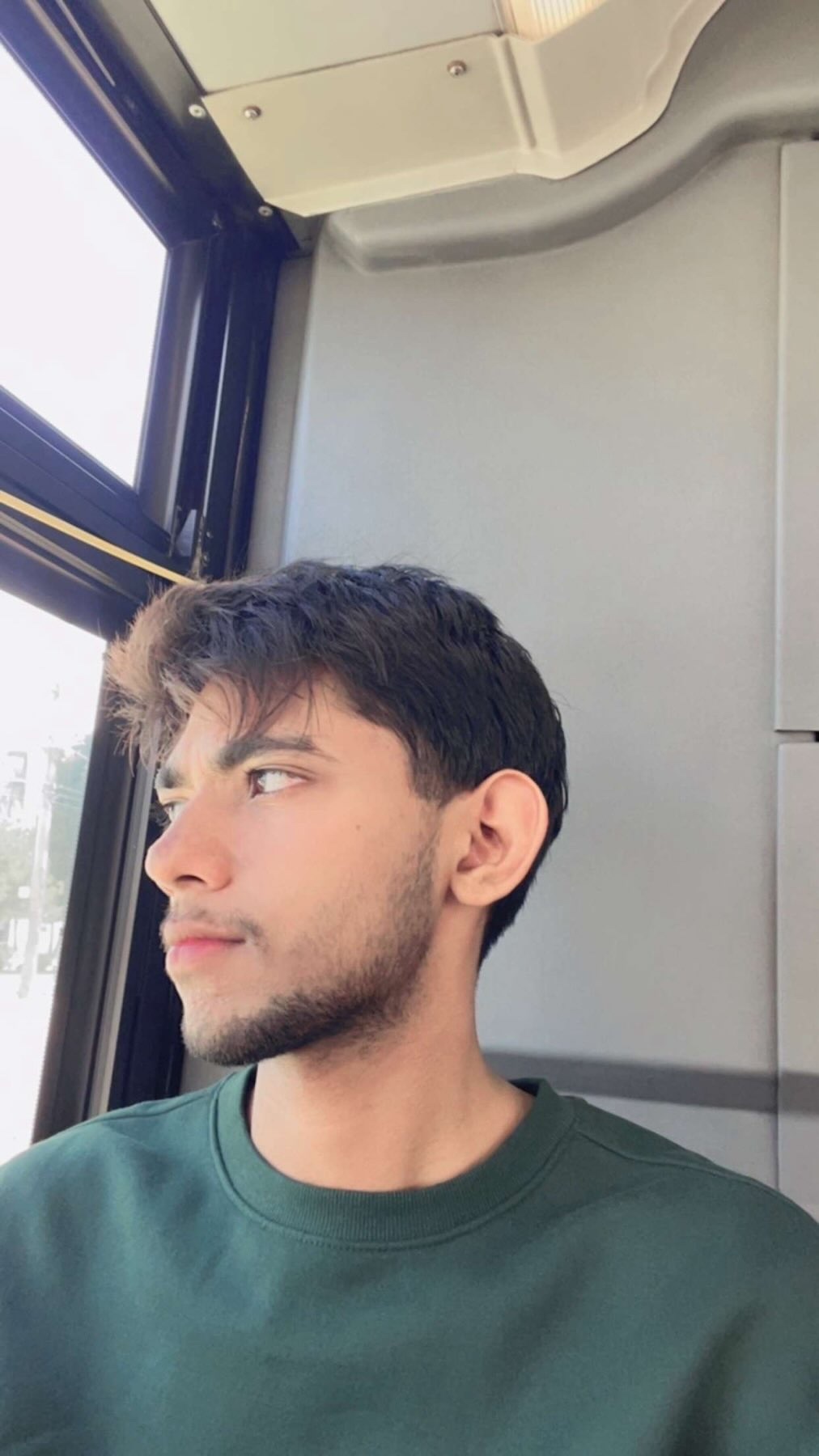 A young man with stubble and dark hair looks out a window while sitting on public transportation.