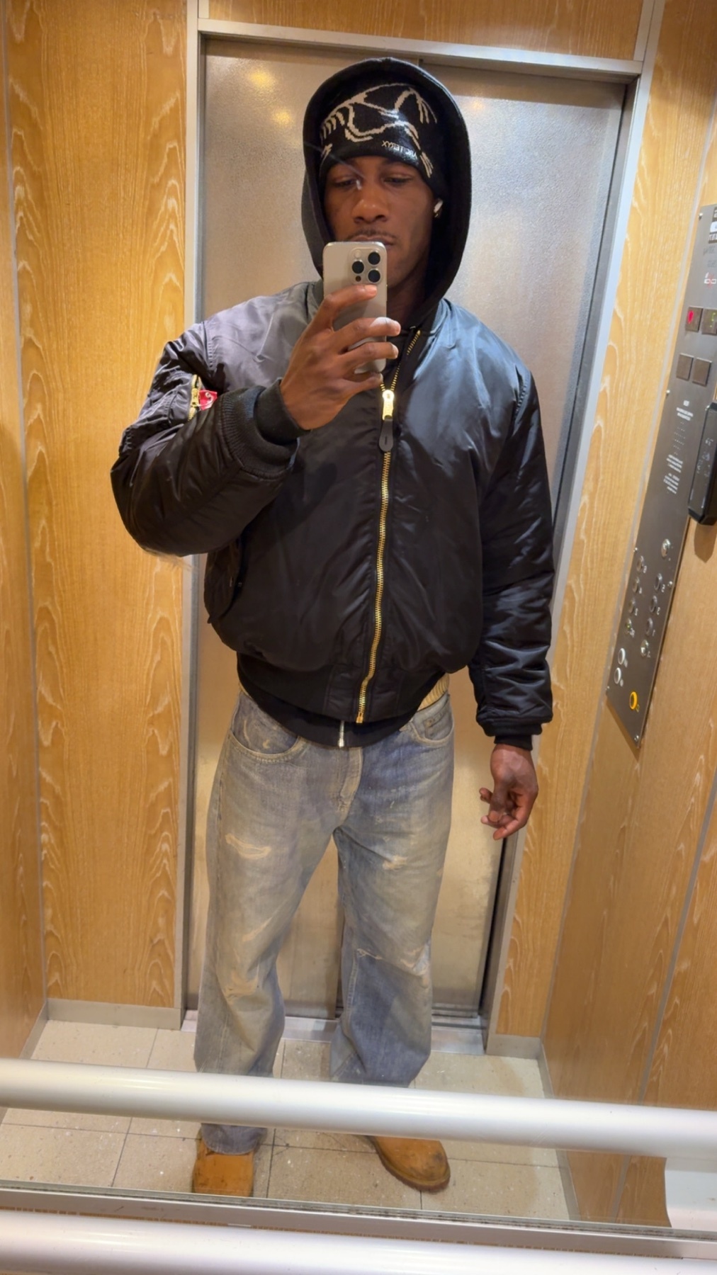 A man takes a mirror selfie in an elevator. He is wearing a black bomber jacket, jeans, and a bandana.