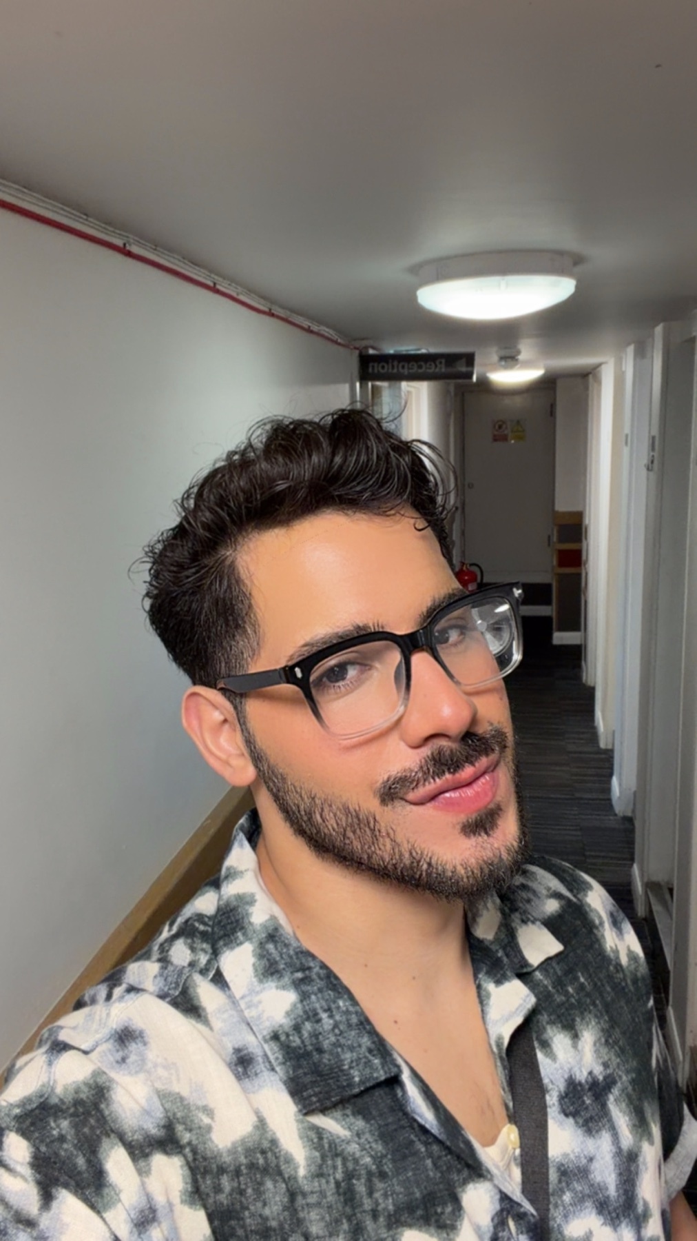 A man with dark, curly hair and a beard poses for a selfie indoors. He is wearing glasses and a black and white patterned shirt.