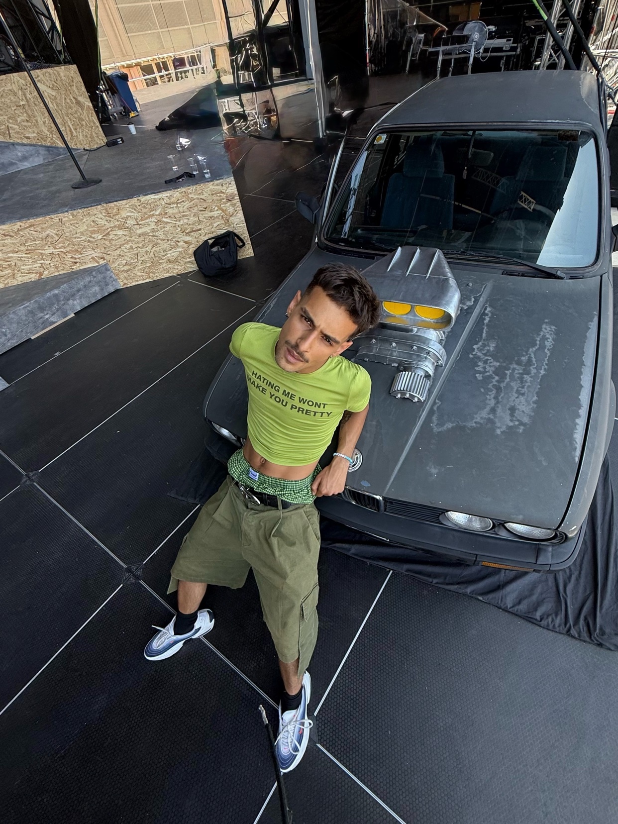 A man poses in front of a car, wearing a green cropped shirt and cargo shorts. The photo is taken from a high angle, showcasing the man's outfit and the car's details.
