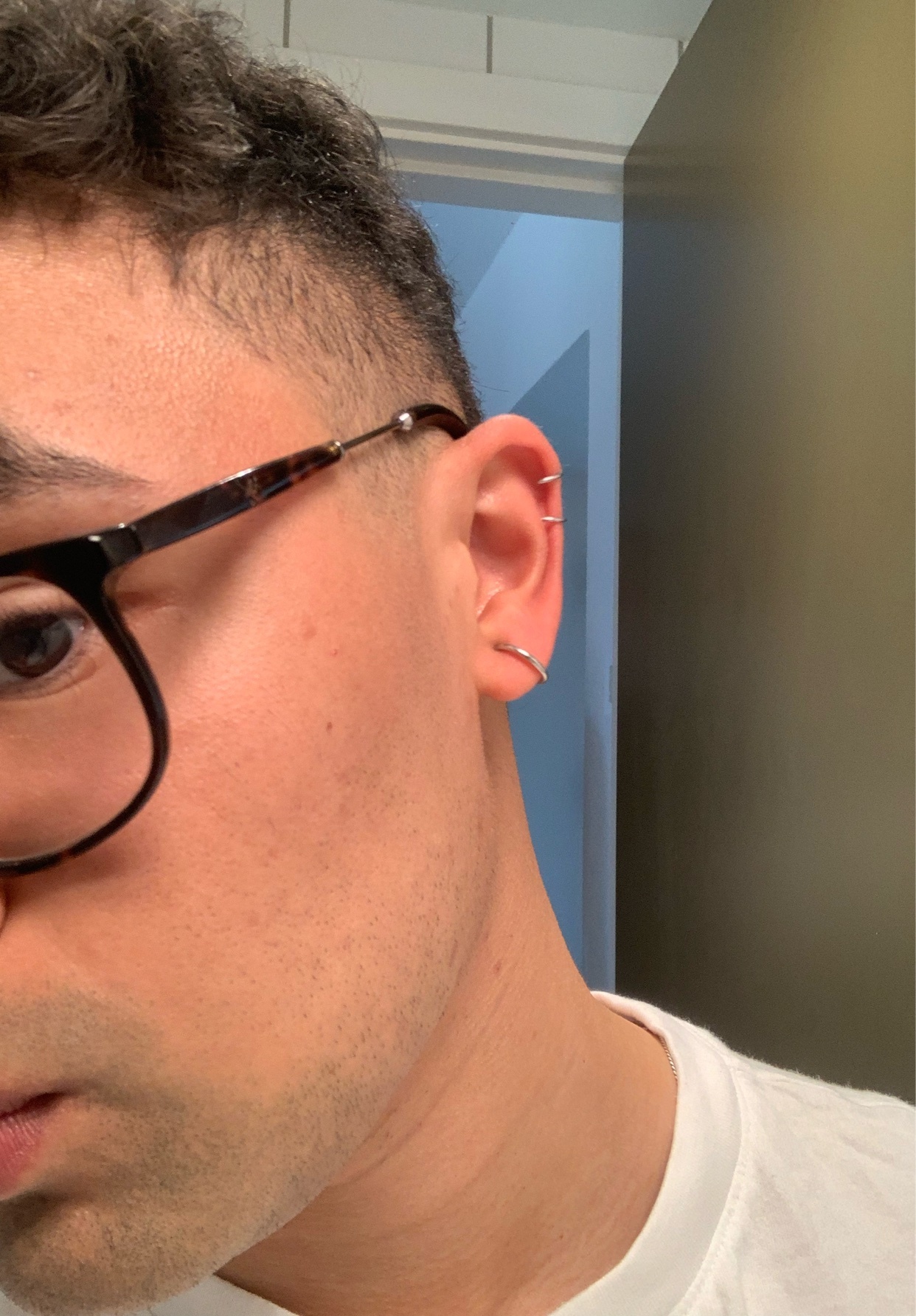A close-up profile shot of a man wearing glasses and multiple earrings in his ear. He is looking towards the camera and appears to be asking for advice.