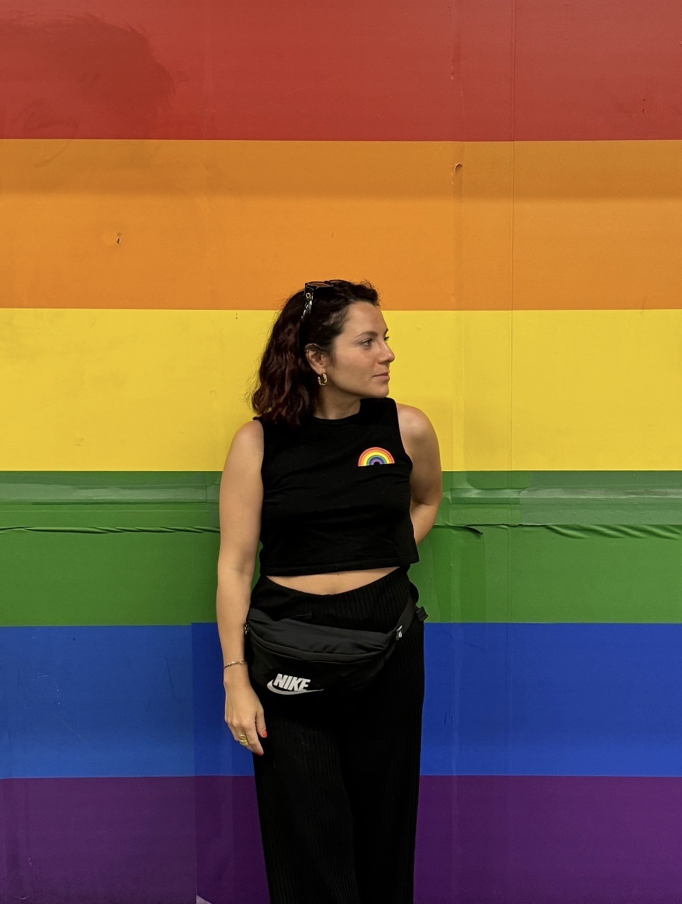 A woman stands in front of a rainbow pride flag wall. She is wearing a black crop top with a small rainbow detail on the chest and a black fanny pack around her waist. The caption is in Italian and translates to 'Here to connect, not to impress. Mayb