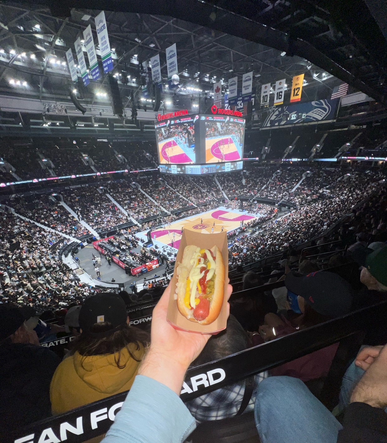 A hand holds a hot dog with mustard, ketchup, and relish in a paper wrapper in front of a crowded basketball arena. The scoreboard shows a game in progress.